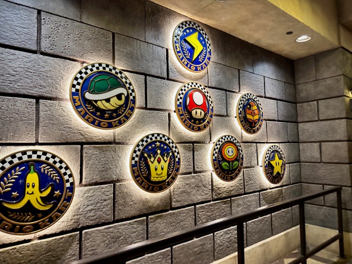 3D badges hanging on a brick wall, with a turtle shell, a banana, a mushroom, a crown, a star, a flower, and a lightning bolt.