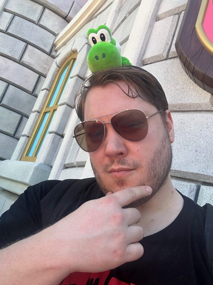 A handsome man with sunglasses and a beard poses with his hand on his chin and a stuffed dinosaur doll on his head.