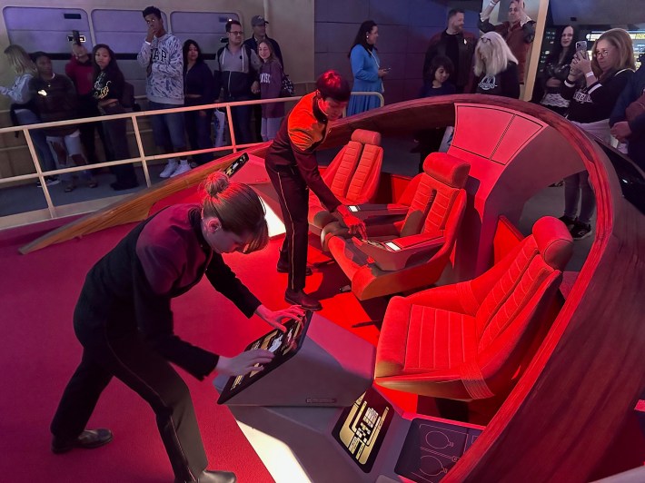 Characters in Star Trek uniforms fiddle with some screens on the bridge of a spaceship.