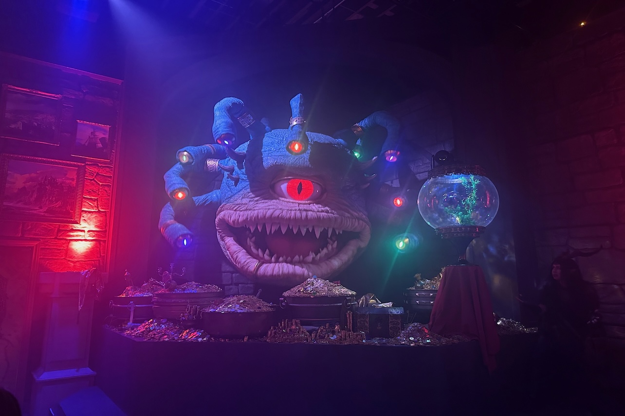 A woman with horns poses next to a large monster puppet, with one eye over its mouth and tentacles.