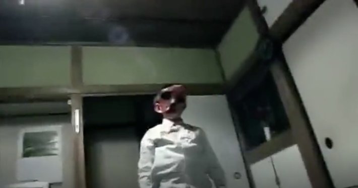A boy with a deformed head stands still in a Japanese living room, faced covered in blood.