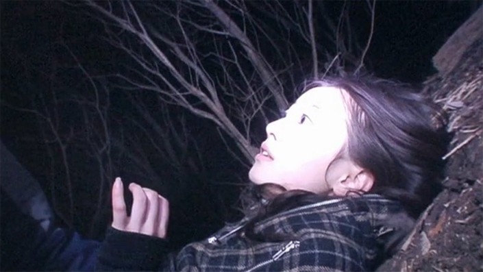 A Japanese woman is lying on her back in the woods, her face illuminated by a TV camera.