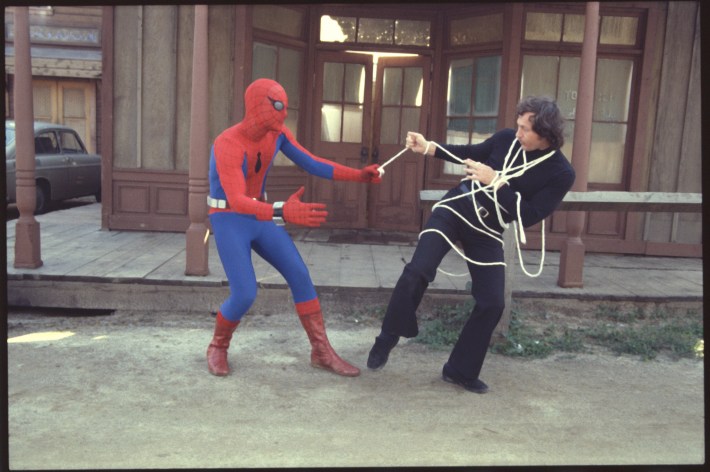 Live action Spider-Man shoots webs out of his hands to entangle a man in a black suit outside a building.