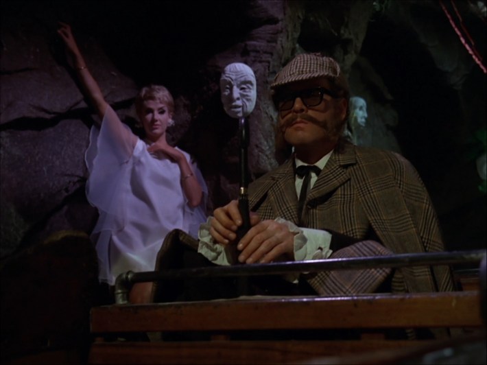A man dressed like Sherlock Holmes is threatened by a woman in a white dress and a grotesque clay mask.