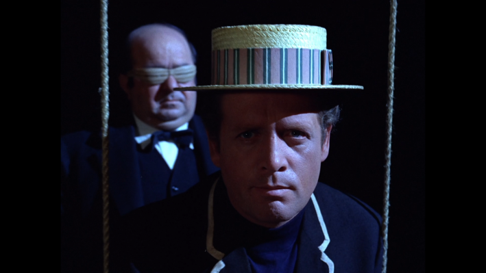 A stern man, Number Six, in a straw hat. Behind him is a masked butler.