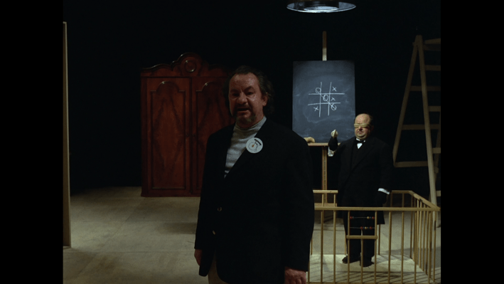 A dark room with a large man presenting evidence, which is a chalk drawing of a tic-tac-toe board, being held by a masked butler.