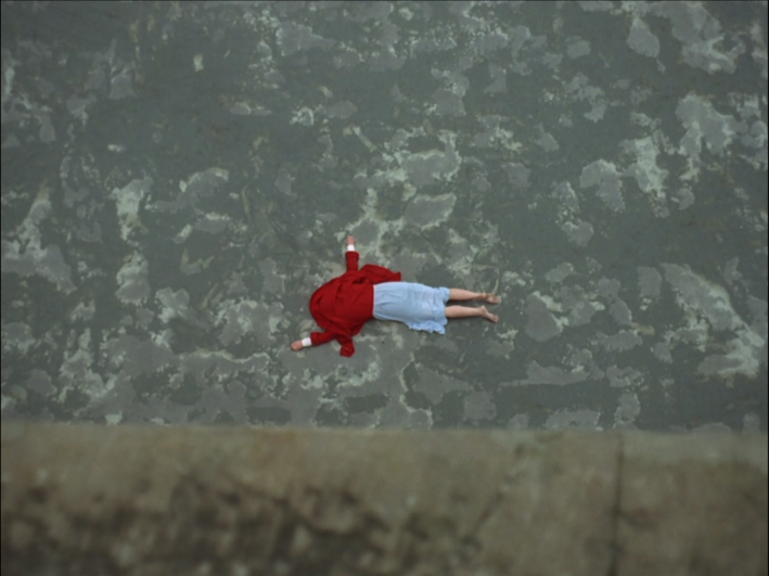 A woman lies face-down on the ground with a red cloak covering her head.
