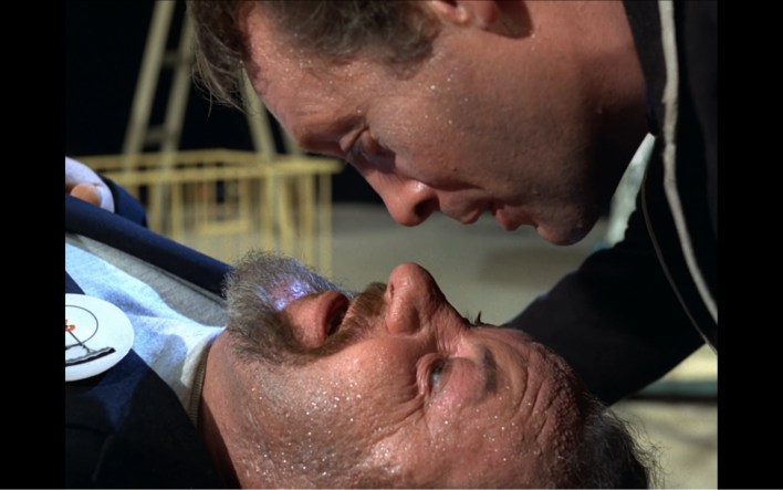 A man, Number Six, peers down at another man, Number Two, who is lying on the floor.