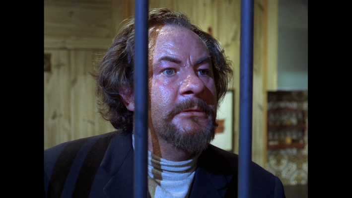 A large bearded man peers out from prison bars.
