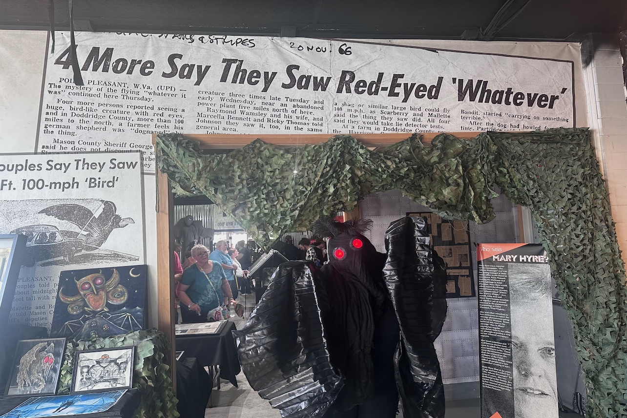 A person dressed in a black wingsuit with red eyes poses underneath a newspaper headline that reads "More say they saw red-eyed 'whatever'".