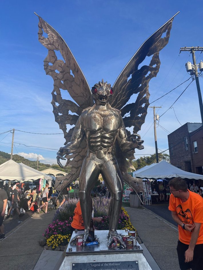 Two people in orange shirts pick up trash by a silver statue of a bug-headed man with wings.