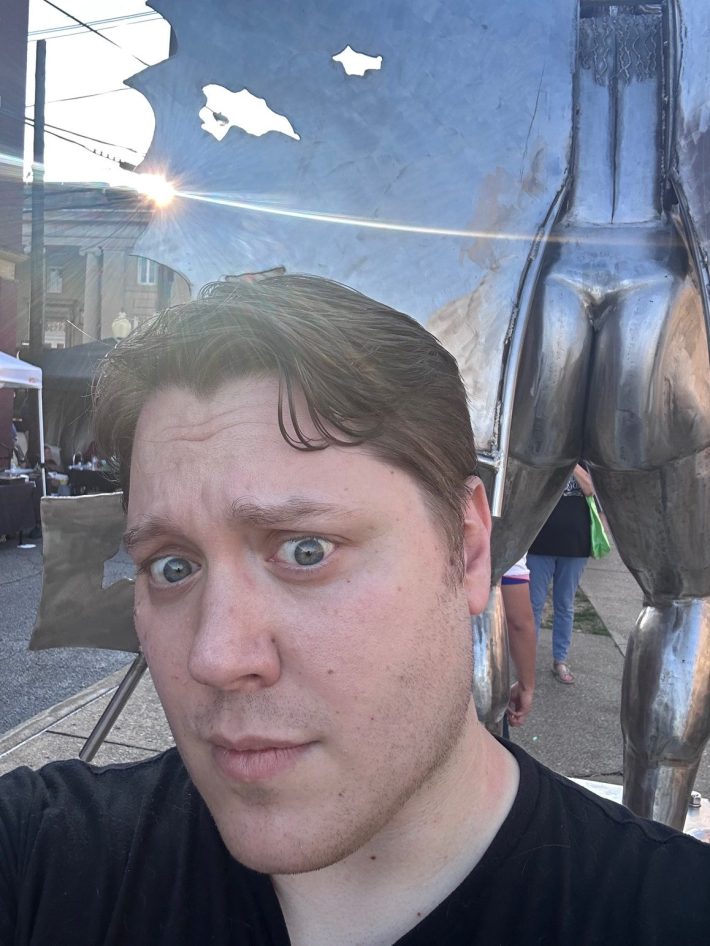 A handsome man cringes next to a silver statue butt.