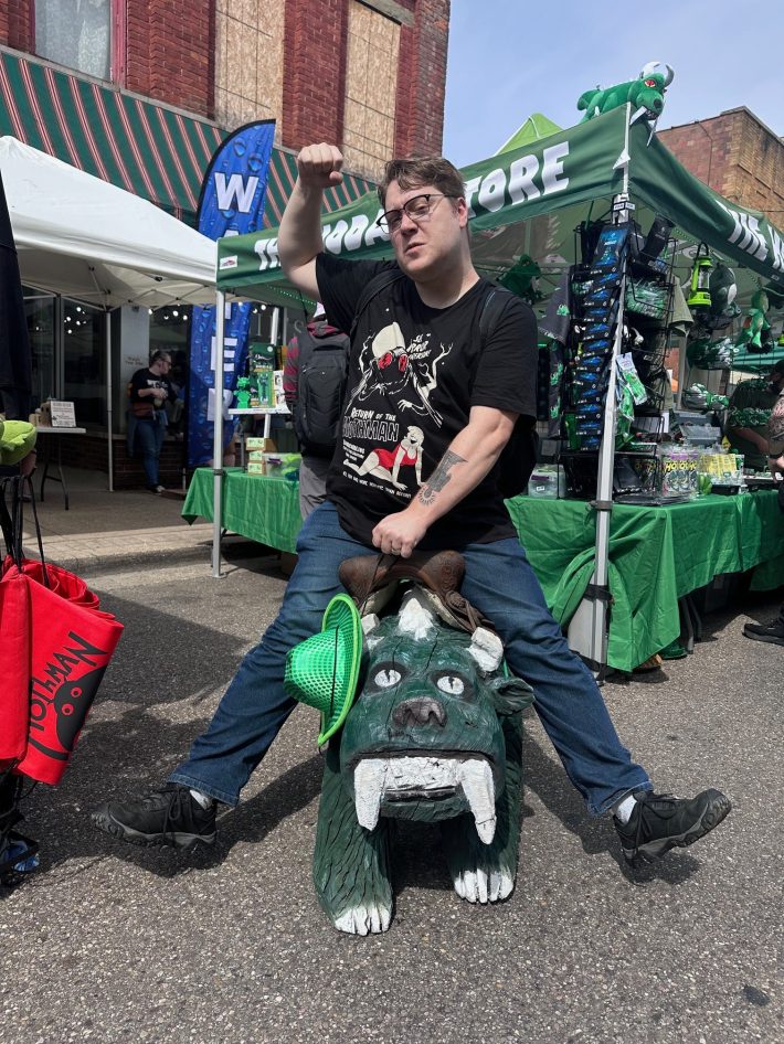 A handsome man poses triumphantly atop a small green monster.