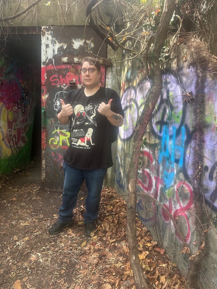 A handsome man poses scared by a concrete bunker covered in graffiti.