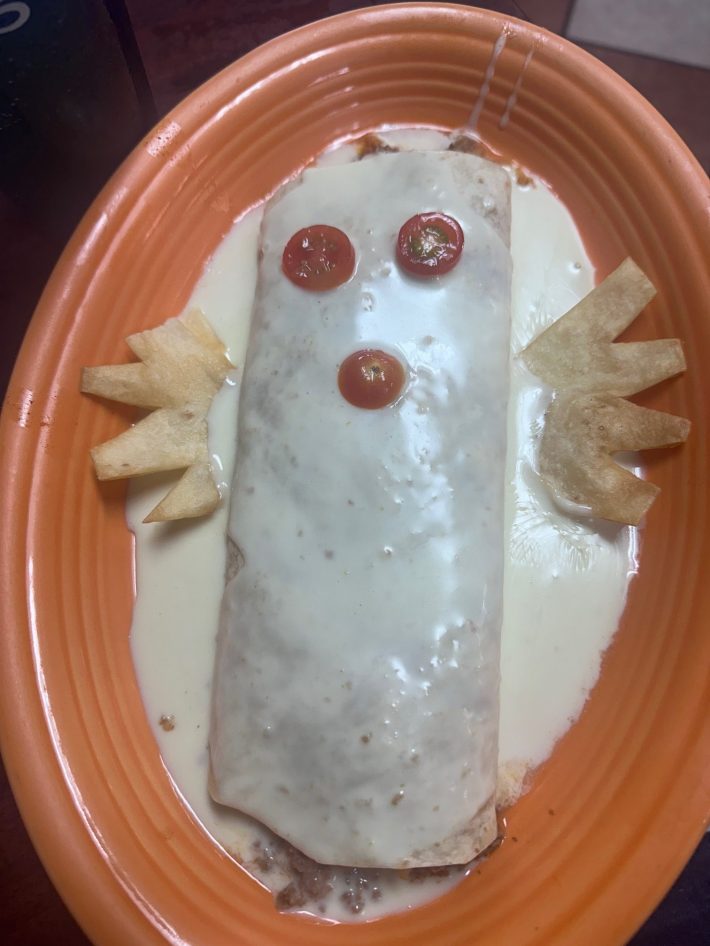 A cheese-covered burrito with tomato eyes and little tortilla wings.