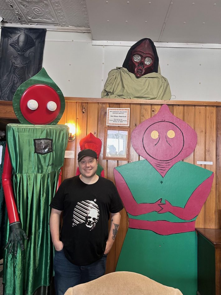 A handsome man poses with three enormous red and green aliens.
