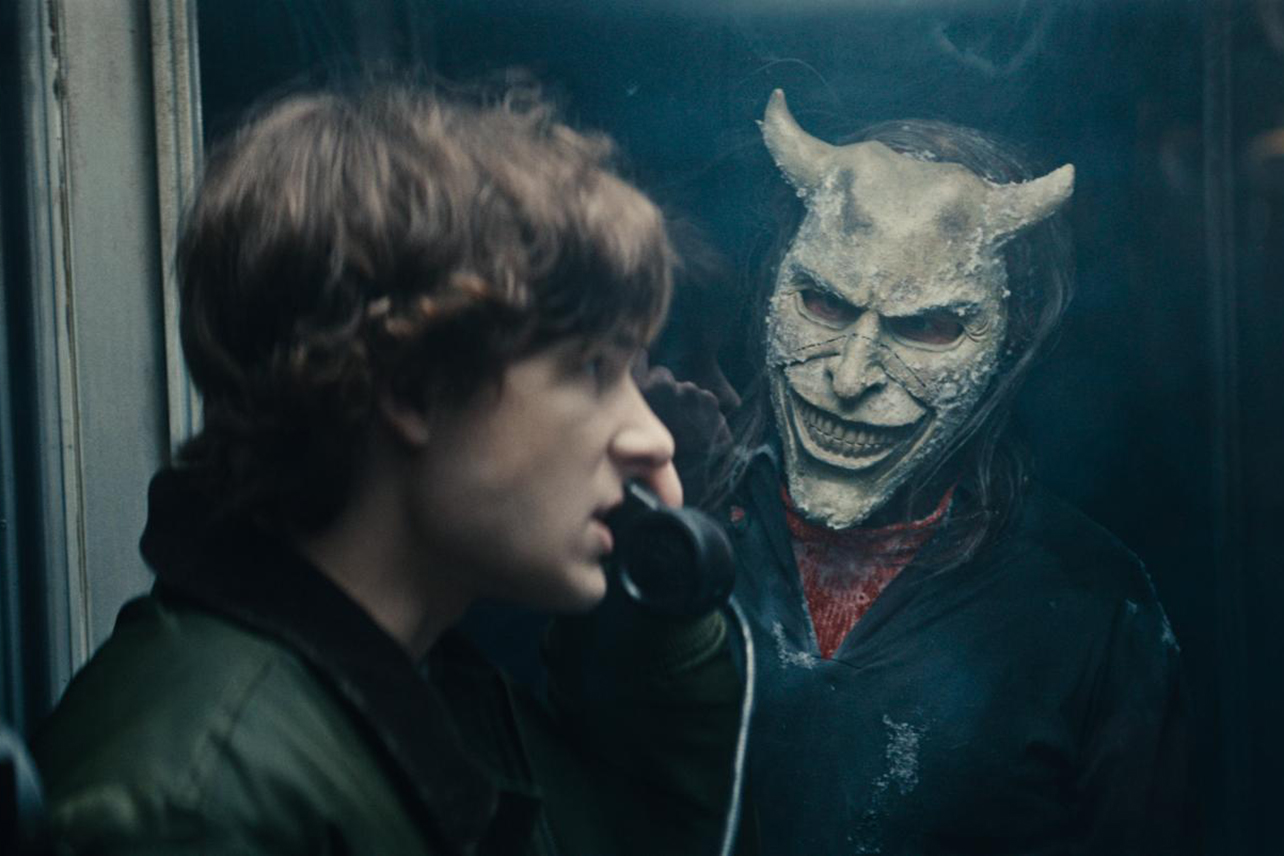 Teen on phone with bloodied man in demon mask looking at him through glass