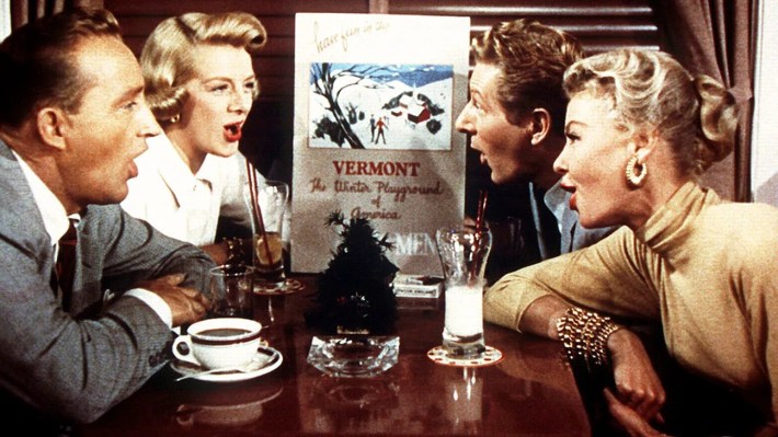 Two men and two women sit at a table singing to a small photo of a ski run.