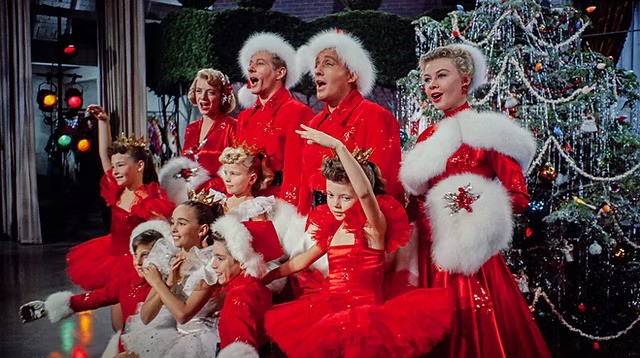 Men and women in red and white furs pose by a Christmas tree by children dancers in red.