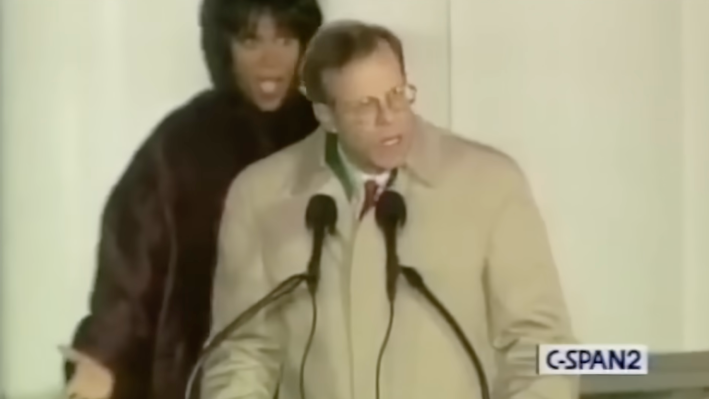 A woman stands behind a man with glasses and a trench coat at a podium. Her mouth is open in shock.