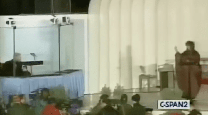 A woman in a burgundy dress waves at a white-haired man far away who is sitting down behind bulletproof glass.