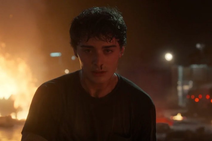 A young man stands in front of an explosion. He is sweaty and dirty, has a pained expression, and a small trail of blood is coming from one nostril.