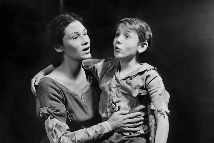 A black and white photo of a peasant mother holding her disabled child. Both of them are singing. The child is holding a crutch.