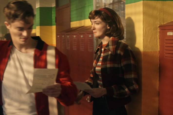 A teenage girl standing in the hallway of a high school as other students walk past.