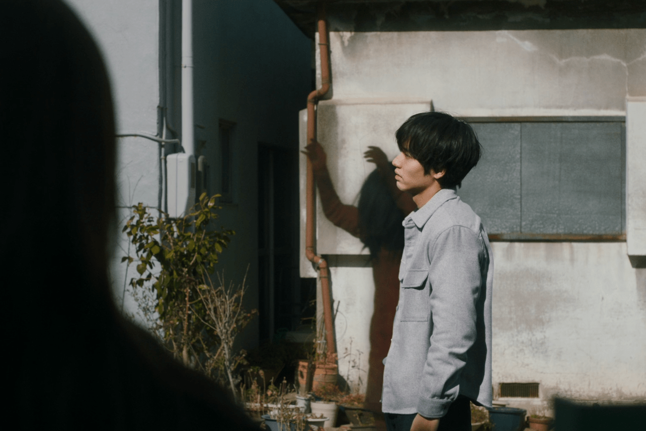 A young Japanese man stands in a trance while a shadow woman stretches her arms to the sky behind him.
